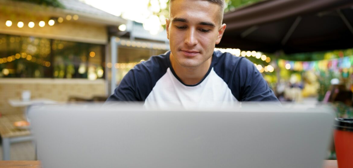 Young man sitting at table and typing on laptop keyboard while working in outdoor cafe