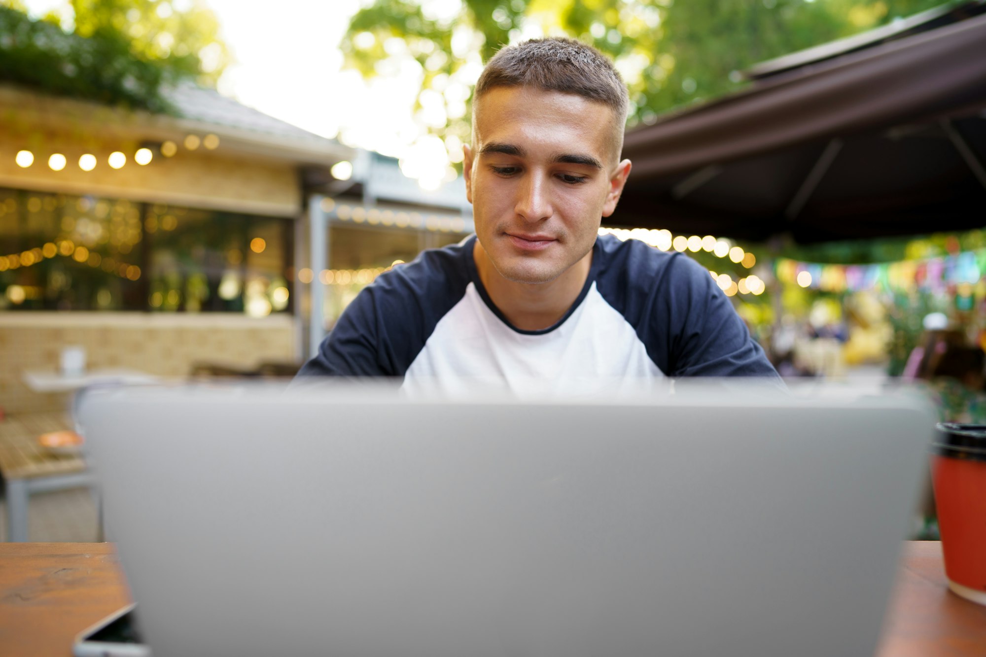 Young man sitting at table and typing on laptop keyboard while working in outdoor cafe